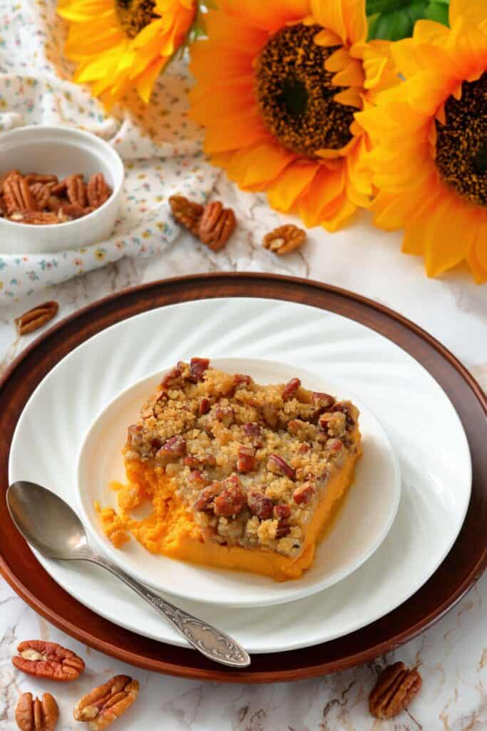 A fully baked sweet potato casserole with a pecan streusel topping, served on a plate with a spoon and surrounded by pecans and sunflowers for a warm, inviting presentation.