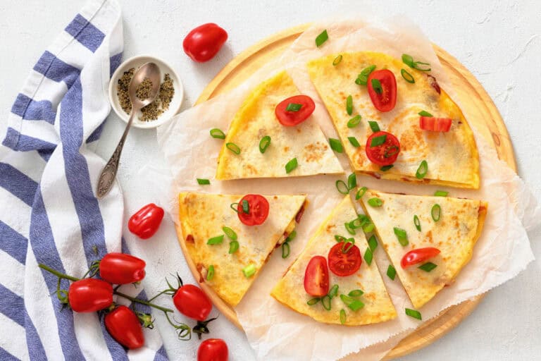 A stack of delicious breakfast quesadillas topped with sliced tomatoes and green onions on a wooden platter, surrounded by fresh cherry tomatoes and a blue striped towel.
