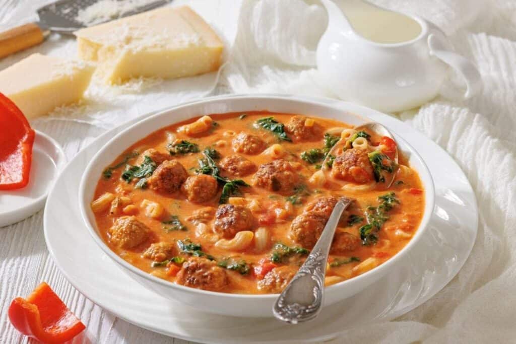 A bowl of meatball soup with pasta, spinach, and tomato broth, served with a spoon.