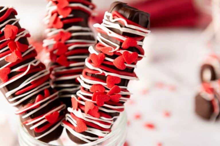Three Valentine’s Day Marshmallow Pops decorated with white icing and red heart-shaped sprinkles stand upright in a glass jar.
