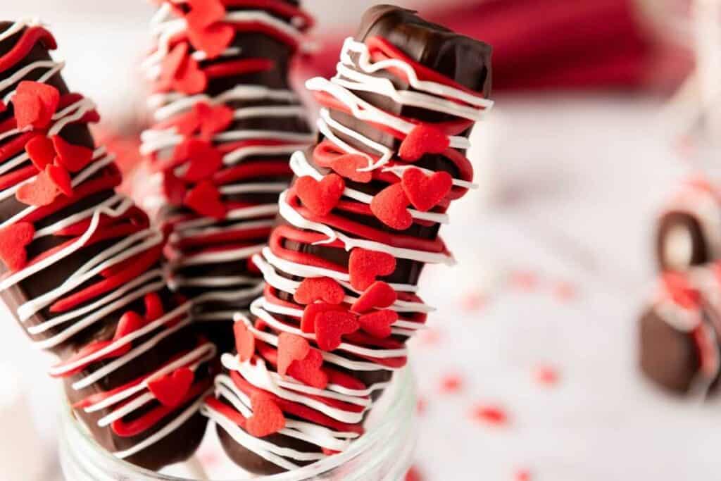 Three Valentine’s Day Marshmallow Pops decorated with white icing and red heart-shaped sprinkles stand upright in a glass jar.