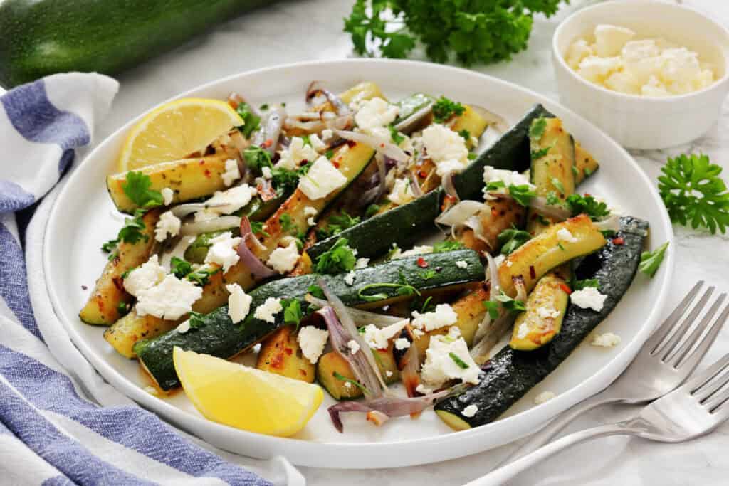 A plate of roasted zucchini and red onions garnished with crumbled cheese, herbs, red pepper flakes, and lemon wedges. A small bowl of cheese and fresh parsley are in the background.