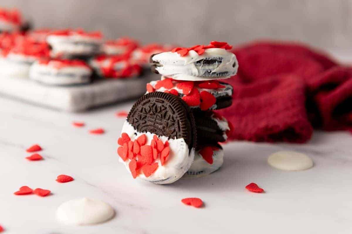 Valentine’s Day Oreos decorated with red heart-shaped sprinkles, arranged on a white surface with a red cloth in the background.