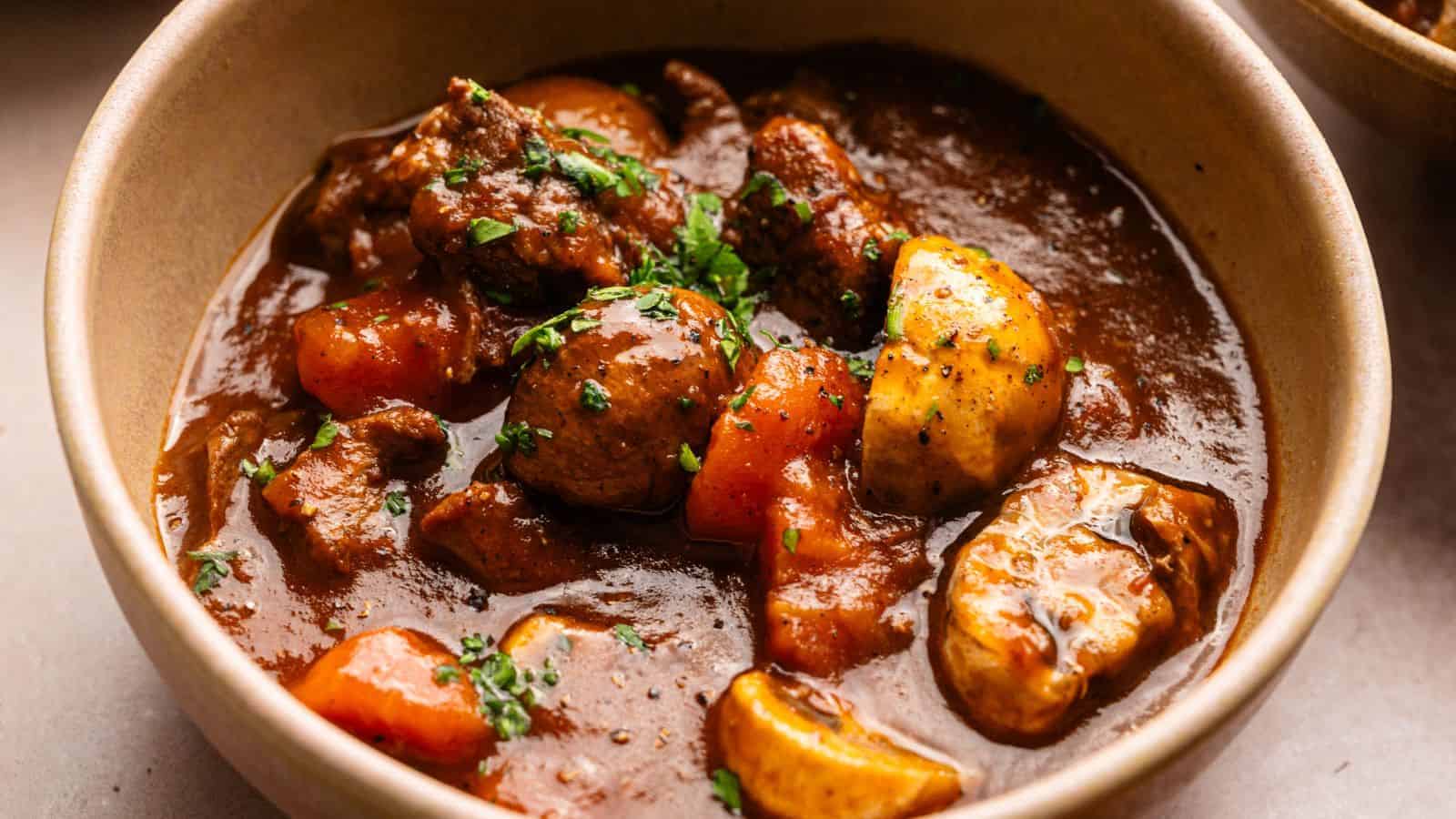 A bowl of beef stew with chunks of beef, carrots, potatoes, and mushrooms in a thick brown broth, garnished with chopped herbs.