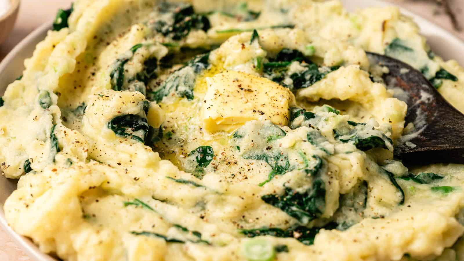A bowl of mashed potatoes with spinach, topped with a pat of butter and black pepper, with a wooden spoon partially visible.