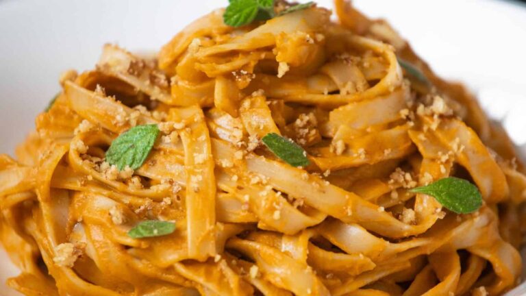 A close-up of fettuccine pasta coated in a creamy orange sauce, garnished with chopped nuts and fresh green herbs.