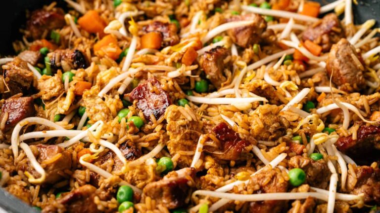 A dish of fried rice featuring chunks of meat, scrambled egg, green peas, diced carrots, and bean sprouts, all stir-fried to a rich brown color and served in a close-up presentation.