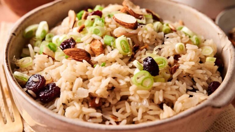 A bowl of rice garnished with sliced green onions, whole and chopped almonds, and dried cranberries, showcasing a colorful and textured dish with a mix of savory and sweet elements.