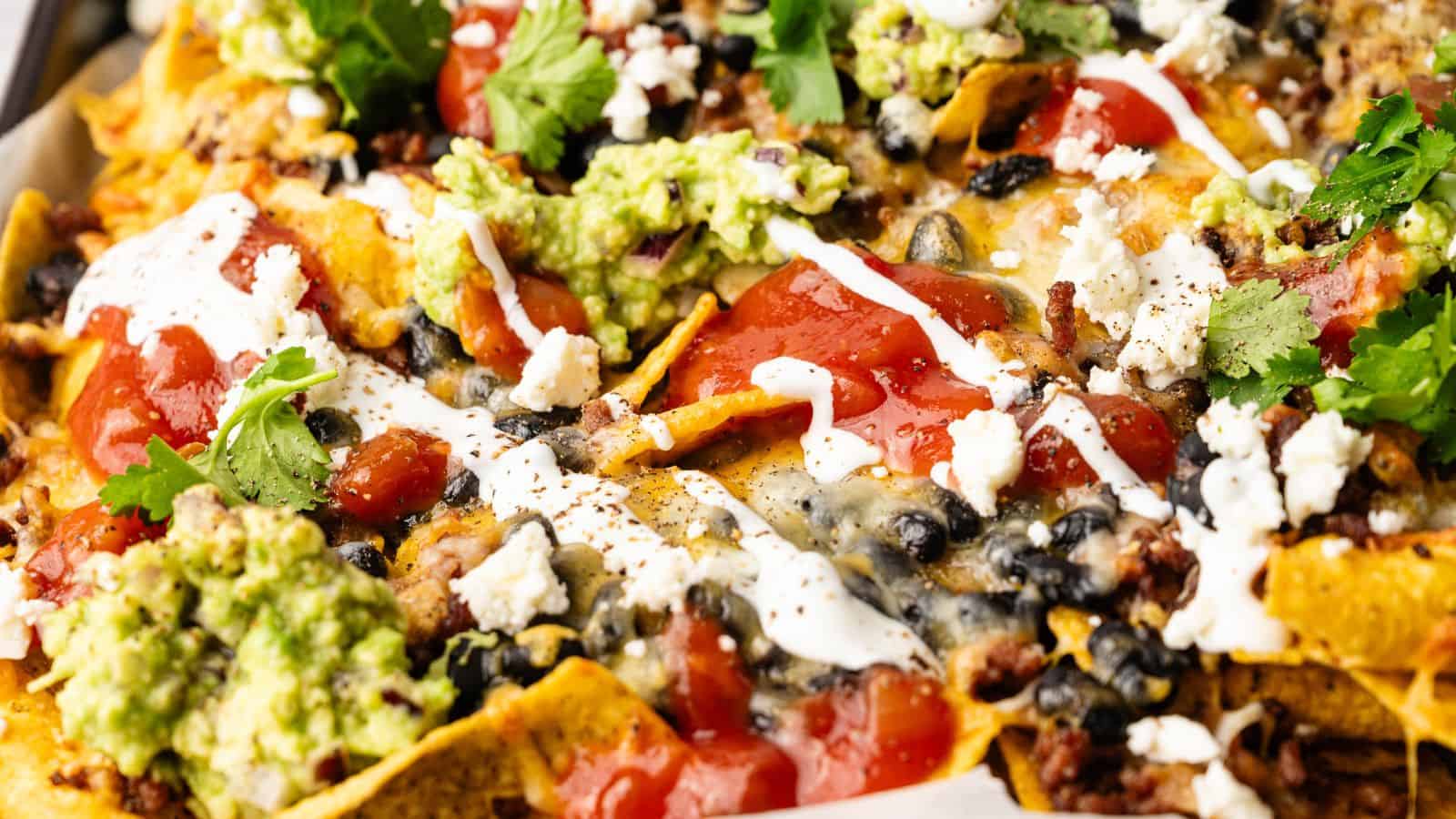 Close-up of nachos topped with melted cheese, black beans, ground meat, guacamole, salsa, sour cream, and fresh cilantro.