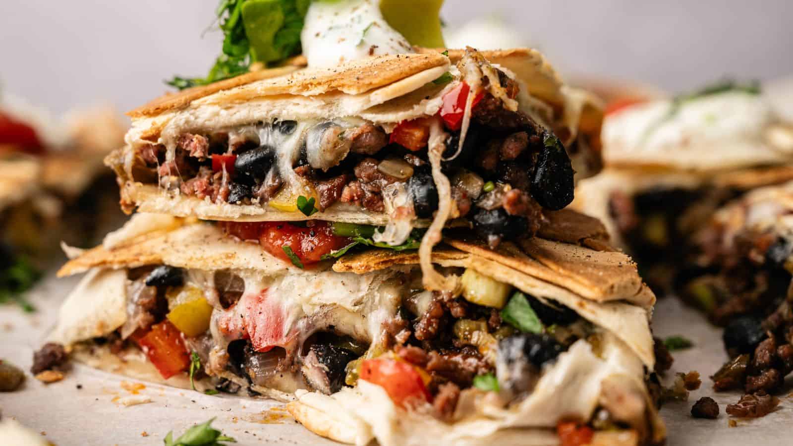 A close-up of stacked quesadilla wedges filled with black beans, ground meat, melted cheese, and diced vegetables, topped with sour cream, guacamole, and chopped herbs.