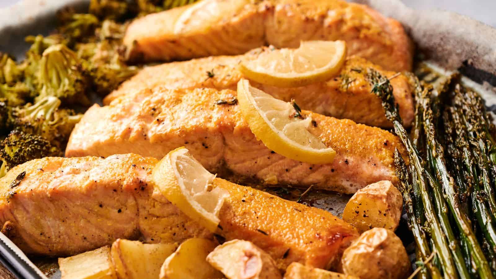 Baked salmon fillets garnished with lemon slices, served with roasted broccoli, asparagus, potatoes, and garlic cloves on a sheet pan.