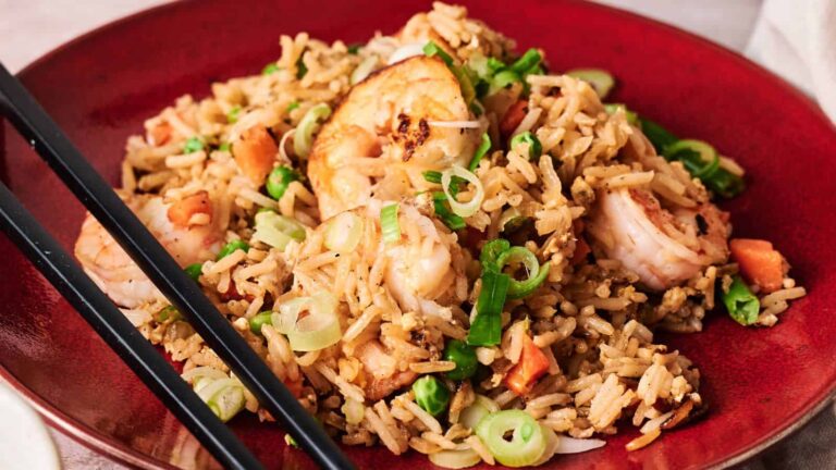 A plate of shrimp fried rice served on a red dish, featuring golden-brown rice stir-fried with shrimp, green peas, diced carrots, chopped green onions, and scrambled egg, with black chopsticks resting on the edge of the plate.