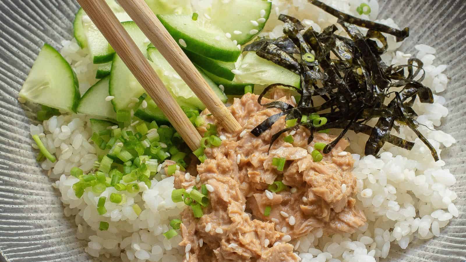 A bowl of white rice topped with sliced cucumber, shredded nori, chopped green onions, and a portion of tuna, with a pair of chopsticks resting on top.