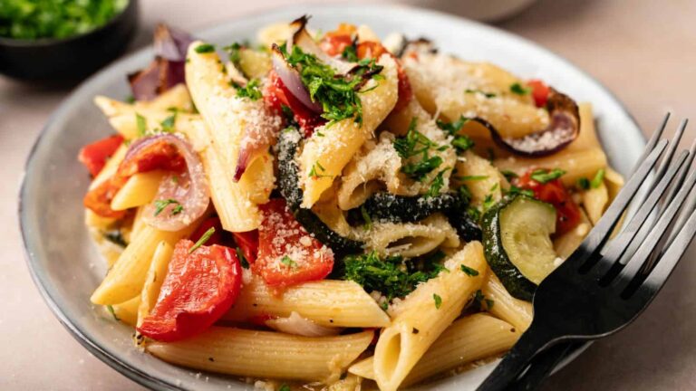A plate of penne pasta mixed with roasted zucchini, red bell peppers, and red onions, garnished with chopped parsley and grated Parmesan cheese, with a black fork resting on the side.