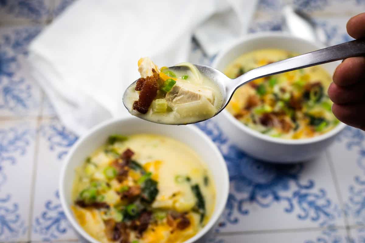 A close-up of a spoonful of Million Dollar soup with chicken, bacon, and green onions held above a bowl of the same soup on a patterned surface.