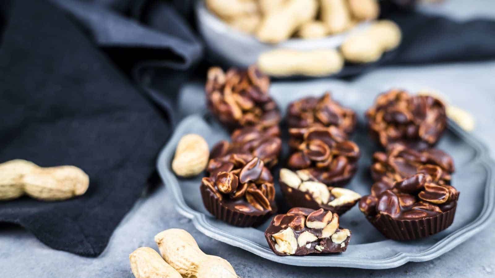 Chocolate peanut clusters on a plate with whole peanuts scattered around on a gray surface.