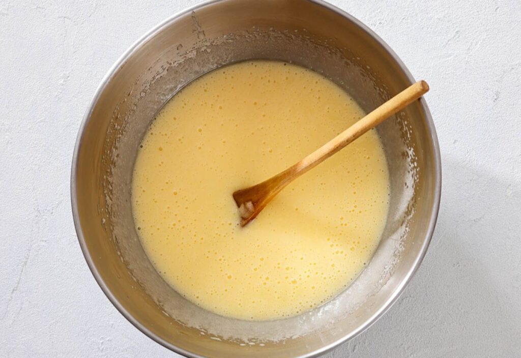 Stainless steel bowl with smooth pale yellow batter stirred by a wooden spoon.