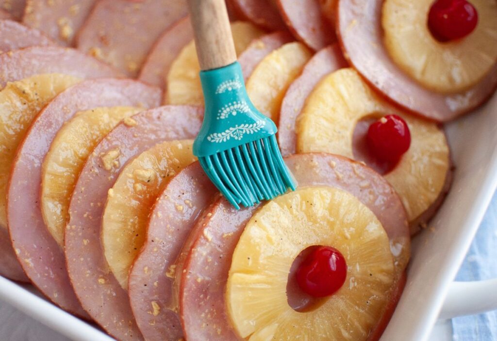 Closeup shot of brushing of glaze on top of layered ham slices with pineapple rings and cherries.
