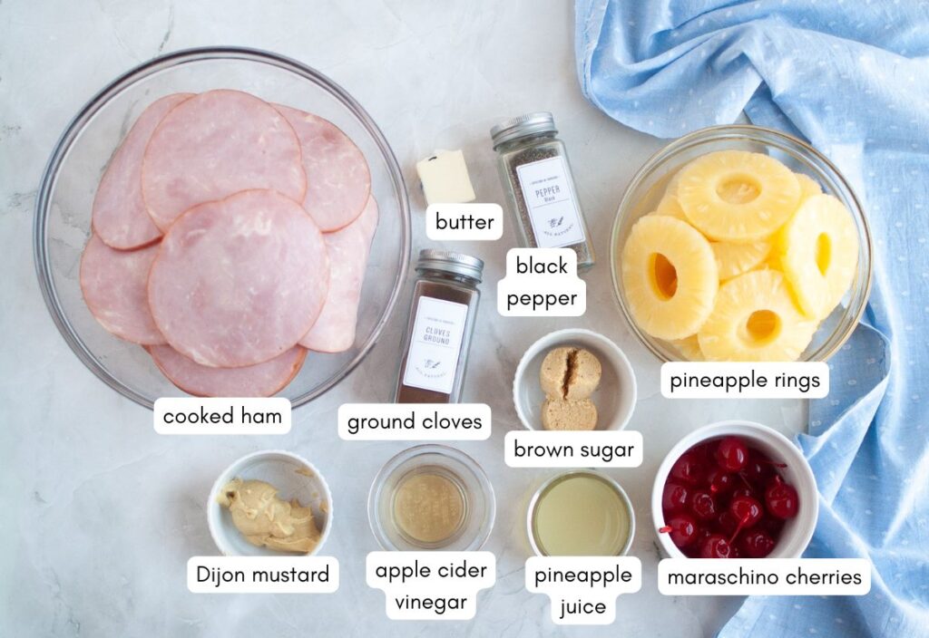 Top view of ingredients needed to create glazed ham slices, including cooked ham, pineapple rings, maraschino cherries, butter, black pepper, ground cloves, brown sugar, Dijon mustard, apple cider vinegar, and pineapple juice.