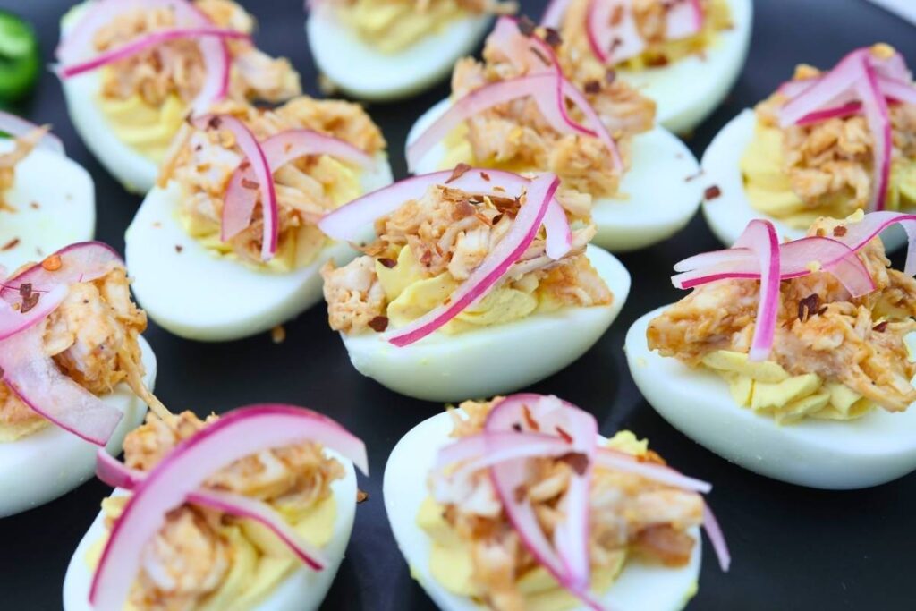 Deviled eggs topped with shredded meat and thinly sliced red onions are arranged on a black plate.