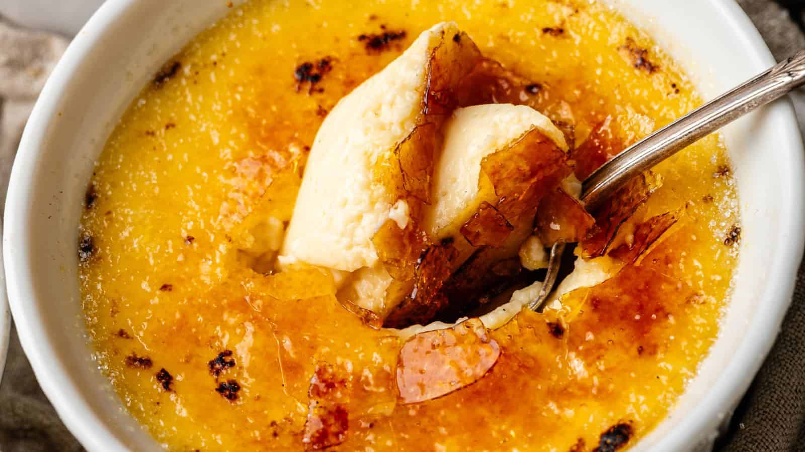 A close-up of crème brûlée in a white dish, showing a caramelized sugar crust being cracked with a spoon to reveal creamy custard underneath.