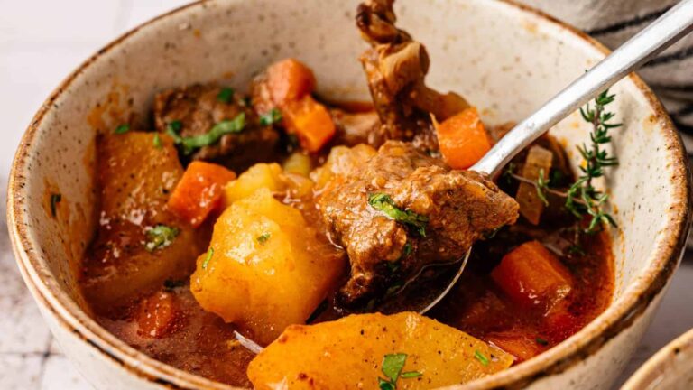 A bowl of beef stew with chunks of beef, potatoes, carrots, and herbs in a rich broth, with a spoon inside the bowl.
