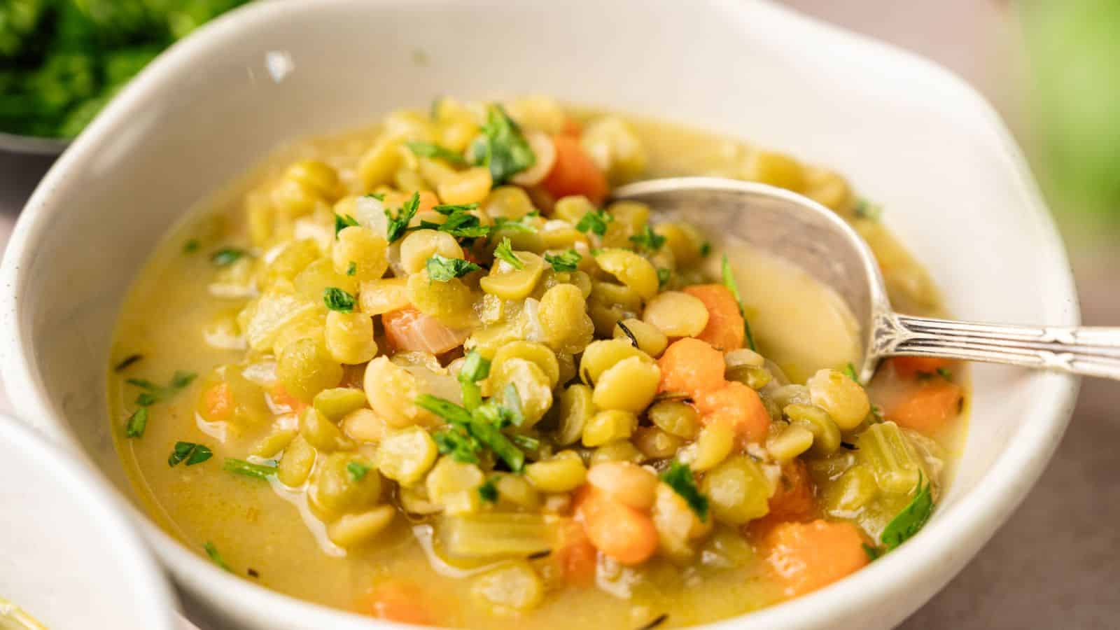 A bowl of split pea soup with carrots, celery, and herbs, with a spoon resting inside the bowl.