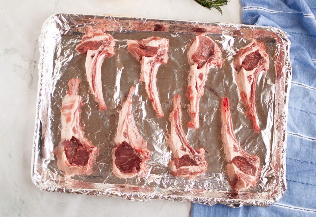 Eight raw lamb chops arranged in a baking sheet lined up with foil.