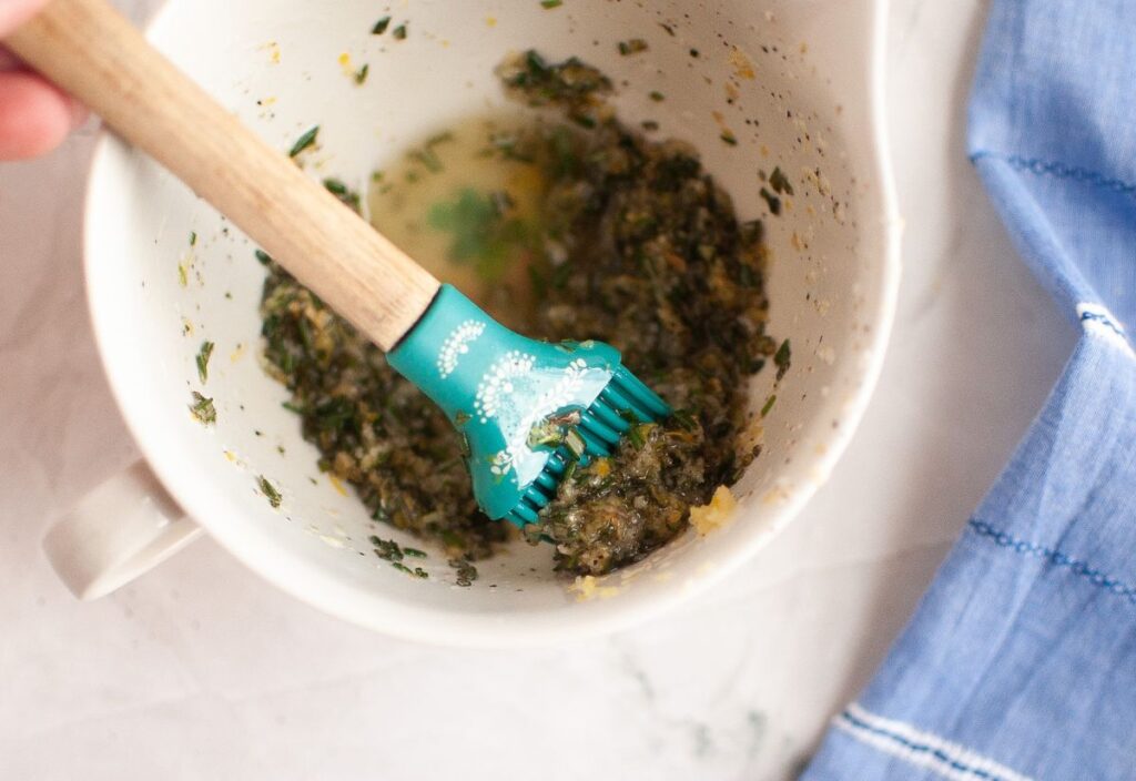 Garlic and rosemary marinade mixed with olive oil, lemon zest, salt, and pepper in a small white bowl with a silicone basting brush.