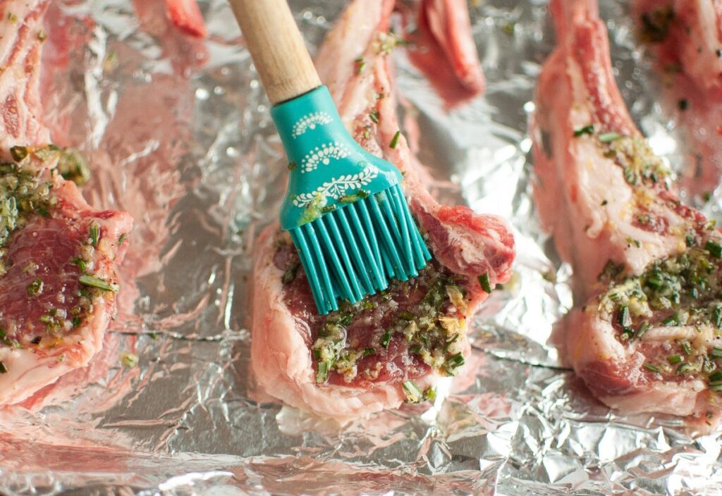 Brushing the garlic and rosemary marinade onto raw lamb chops arranged on a foil-lined baking sheet with a silicone basting brush.