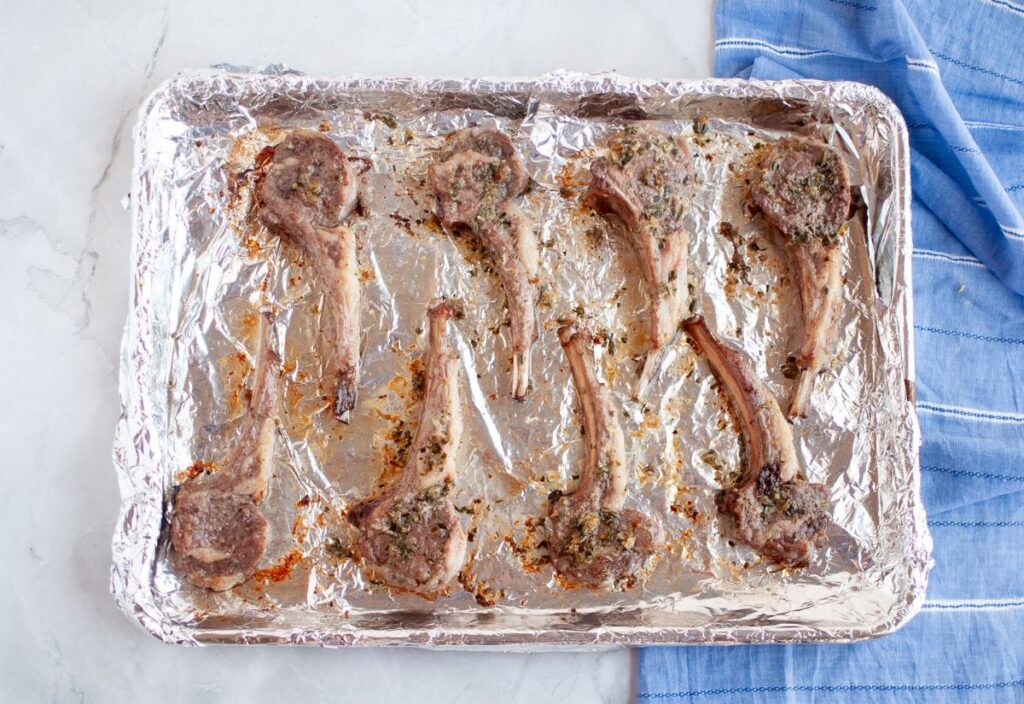Roasted lamb chops with golden brown crusted edges covered in garlic and rosemary on a foil-lined baking sheet after cooking.