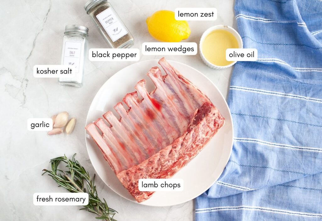 Ingredients for oven roasted lamb chops laid out on a marble surface including raw lamb chops, fresh rosemary, garlic cloves, kosher salt, black pepper, lemon zest, lemon wedges, and olive oil.