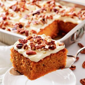 Slice of frosted carrot cake with pecans lifted from a tray.