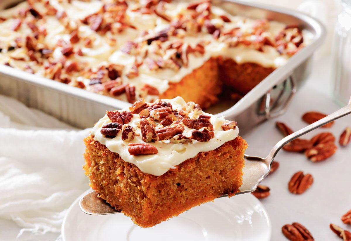 Slice of frosted carrot cake with pecans lifted from a tray.
