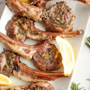 Oven roasted lamb chops with rosemary and garlic arranged on a white plate with fresh lemon wedges and sprigs of rosemary.