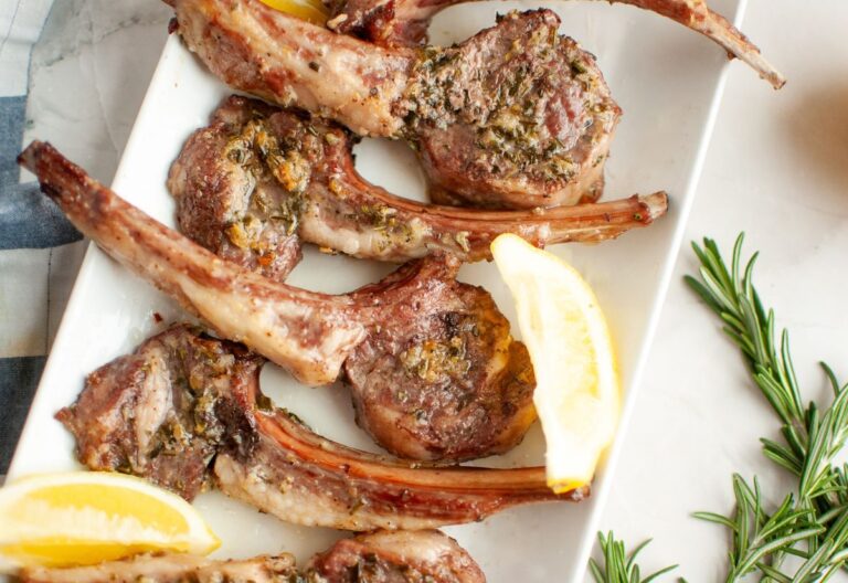 Oven roasted lamb chops with rosemary and garlic arranged on a white plate with fresh lemon wedges and sprigs of rosemary.