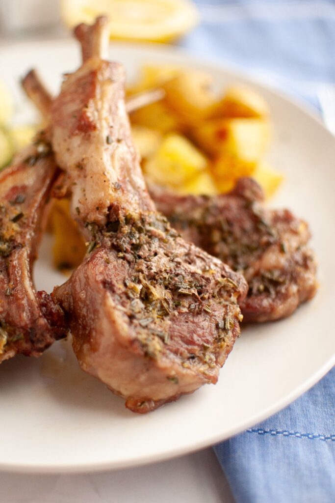 Closeup shot of a plated Oven Roasted Lamb Chops With Rosemary and Garlic with sides on the background.