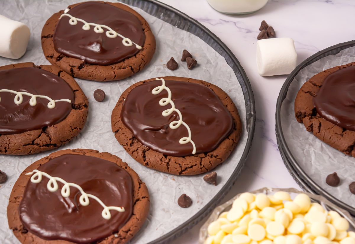 Cookies with a chocolate ganache topping and a white chocolate drizzle, placed on parchment paper with chocolate chips and marshmallows nearby.