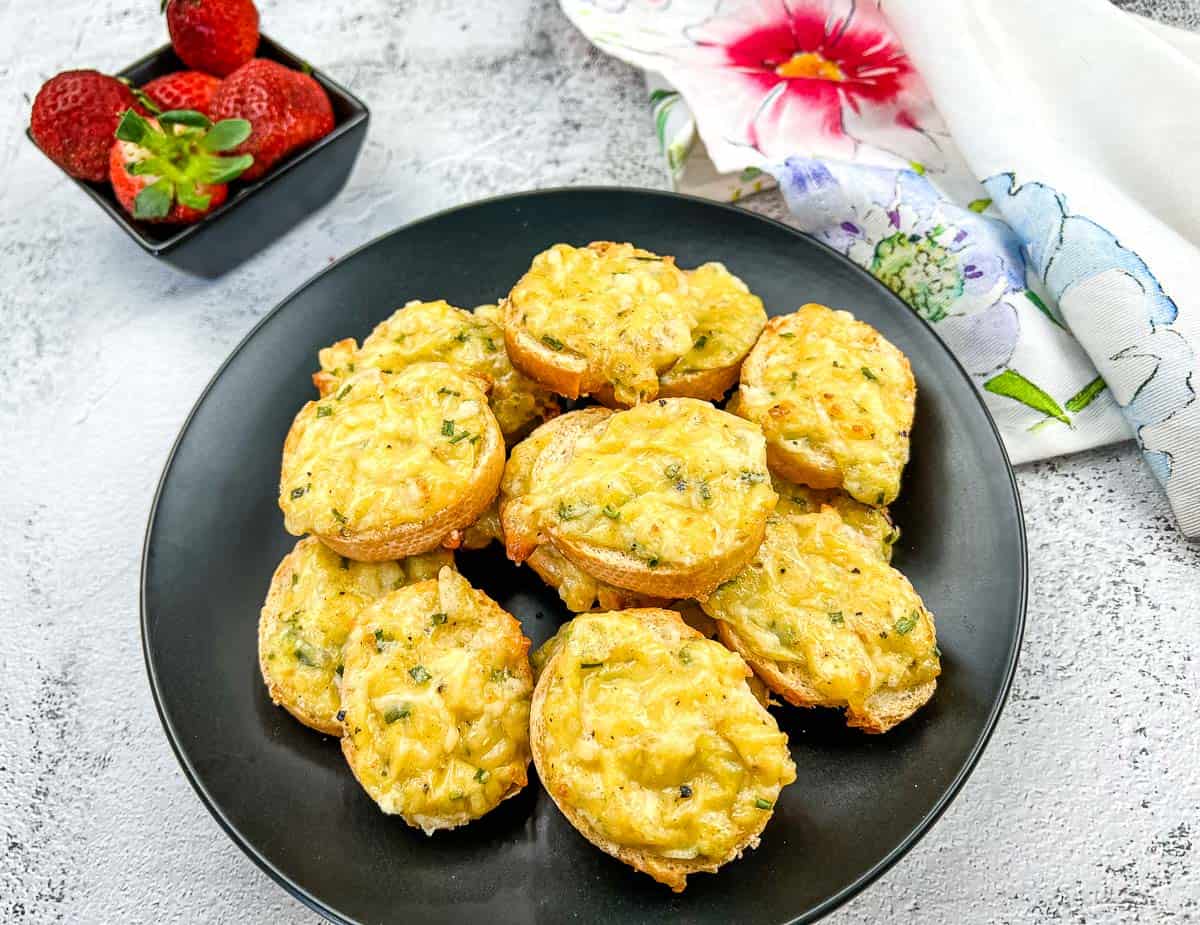 Bite-sized Cheese Toasties on a black plate with a small bowl of strawberries and a floral napkin nearby.