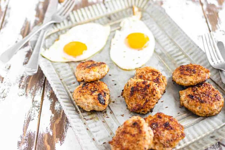 A metal tray holds two sunny-side up eggs and several browned sausage patties. Forks and knives are placed nearby on a rustic wooden surface.