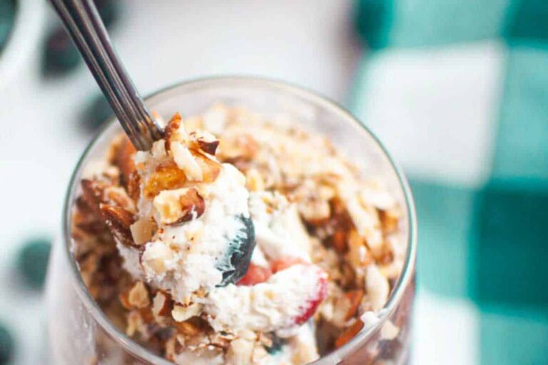 A close-up of a dessert parfait in a glass topped with whipped cream, chopped nuts, and fruit, with a spoon inserted.