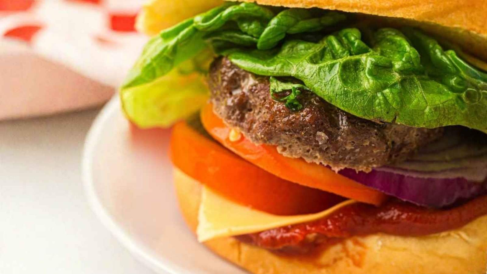A close-up of a hamburger with lettuce, tomato, onion, cheese, ketchup, and a beef patty in a bun, served on a white plate.