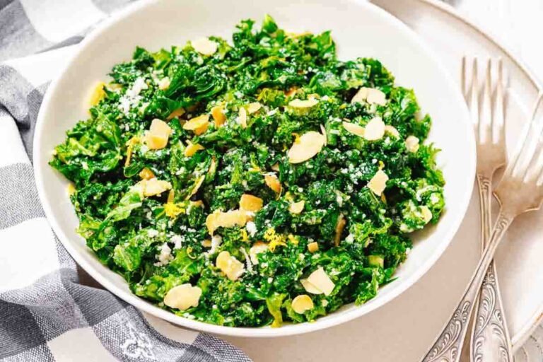A bowl of kale salad topped with sliced almonds and grated cheese, placed on a table with two forks beside it.