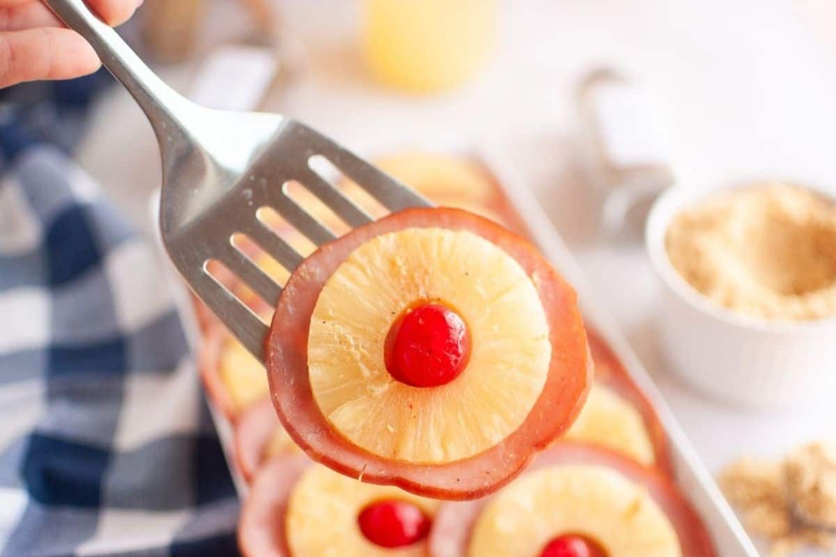 A metal spatula holds a slice of ham topped with a pineapple ring and a red maraschino cherry; more similar slices are visible in the background.