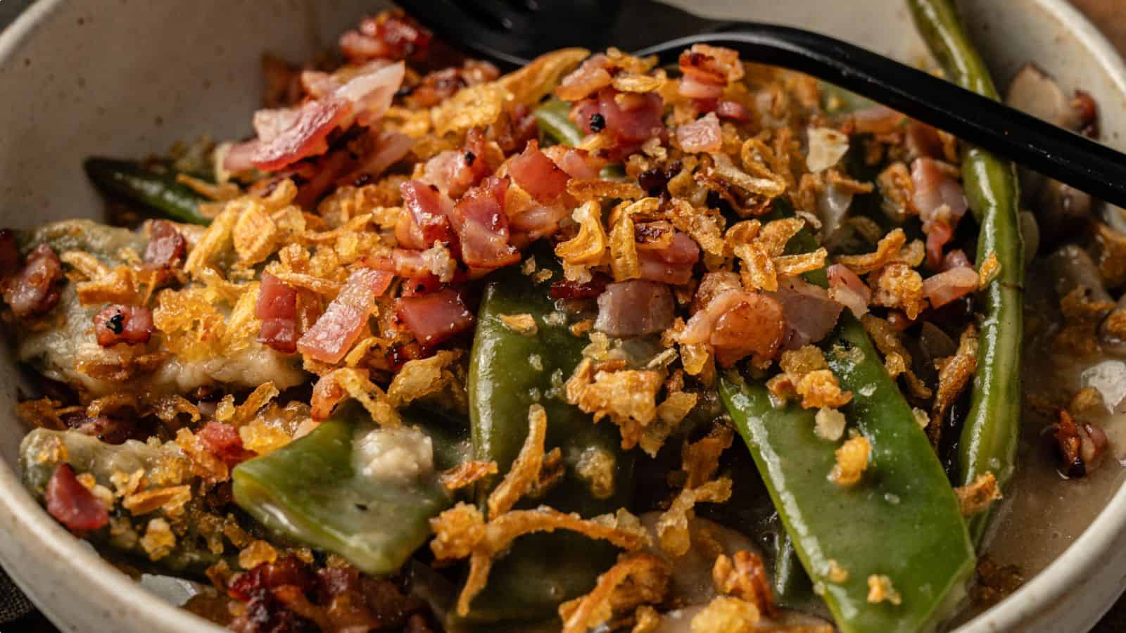 A close-up of a dish with green beans, snap peas, chopped bacon, and crispy fried onions, served in a bowl with a black spoon.