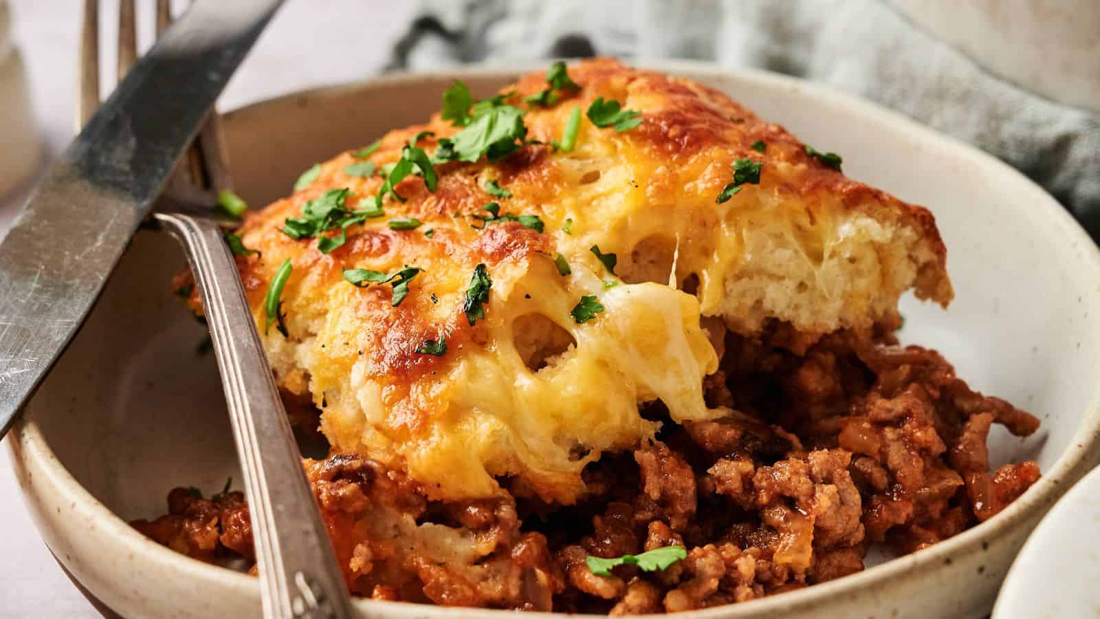 A serving of baked casserole with a golden cheese topping sits on ground meat in a bowl, garnished with chopped parsley and accompanied by a fork and knife.