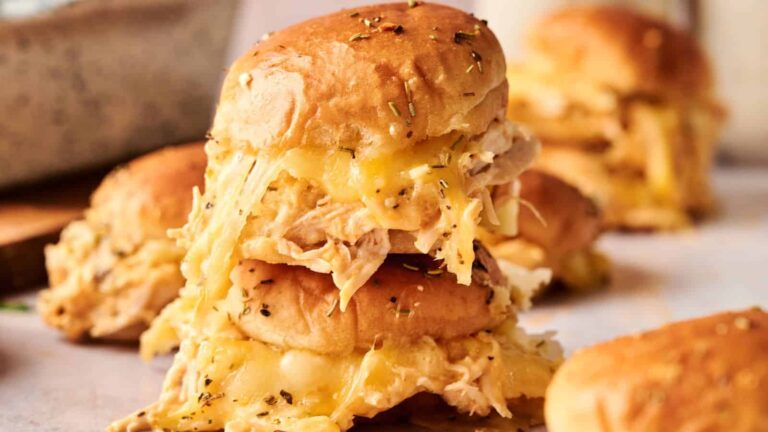 Two stacked cheesy chicken sliders with melted cheese and herbs on soft buns, surrounded by similar sliders on a light surface.
