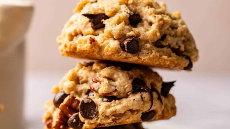 Three chocolate chip cookies are stacked on top of each other with a blurred mug in the background.