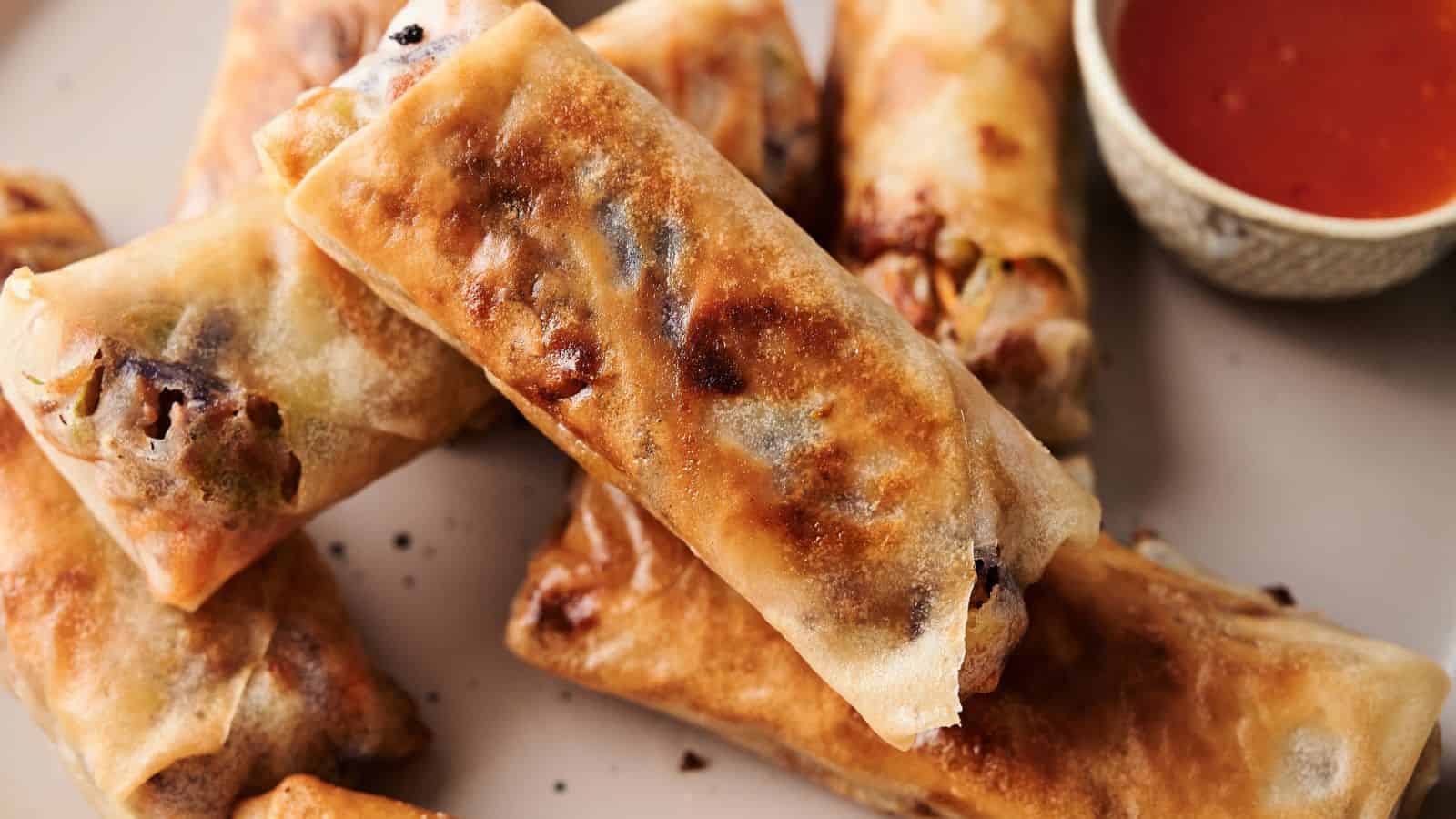 A wooden tray with spring rolls and lime wedges lined with parchment paper sits next to a small bowl of dipping sauce on a light surface.