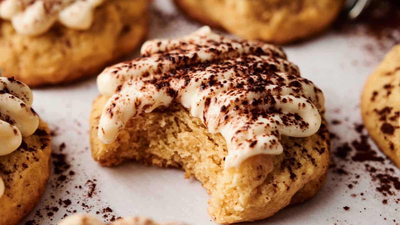 A soft cookie topped with cream and cocoa powder has a bite taken out of it. Other similar cookies are visible in the background.